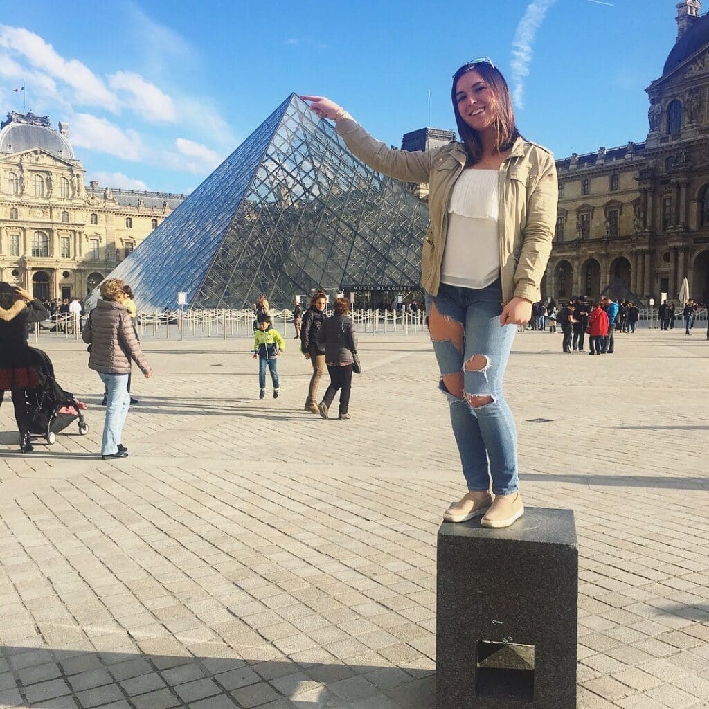 Nadia Filipic on square in front of The Louvre pointing to the tip of the glass pyramid